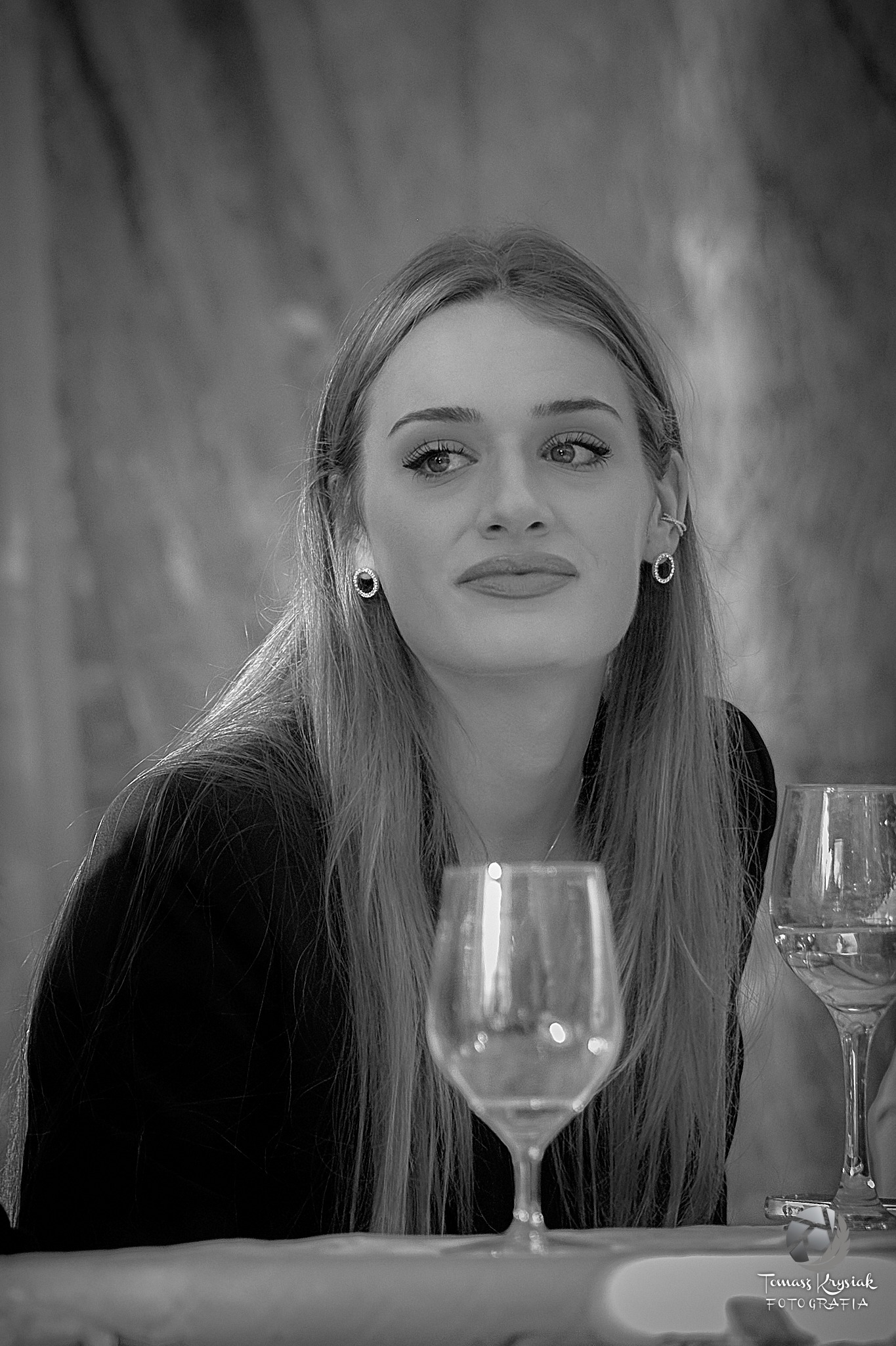 Czarno-biały portret młodej kobiety z długimi włosami, noszącej kolczyki, z kieliszkami w tle, spoglądającej w lewo.
