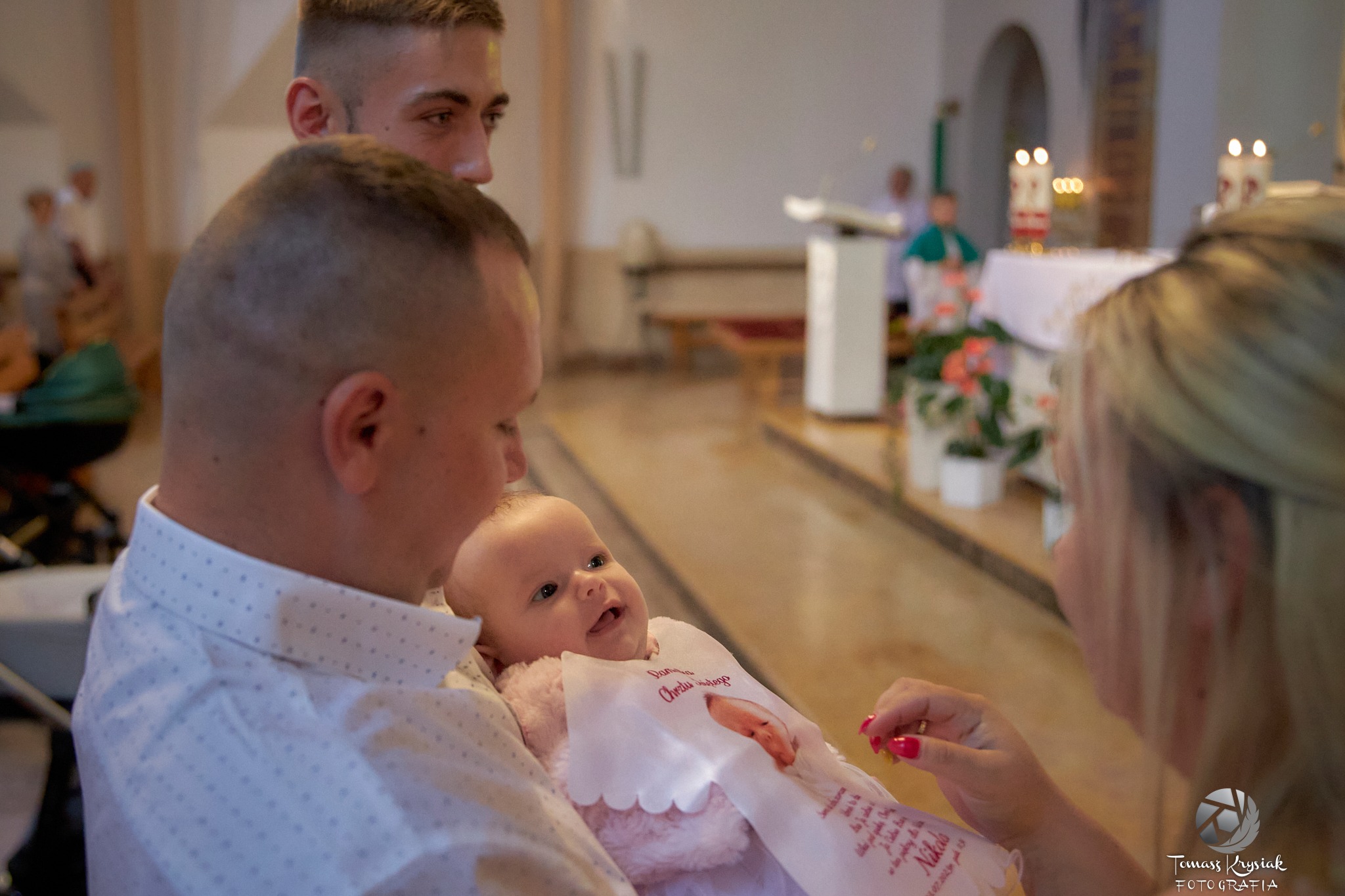 Uśmiechnięte niemowlę w ramionach ojca podczas ceremonii chrztu w kościele, w tle świece i osoby