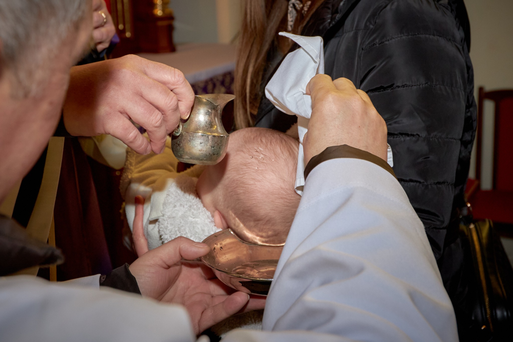 Ksiądz polewa wodą głowę niemowlęcia podczas chrztu, w tle widoczna osoba w czarnej kurtce.