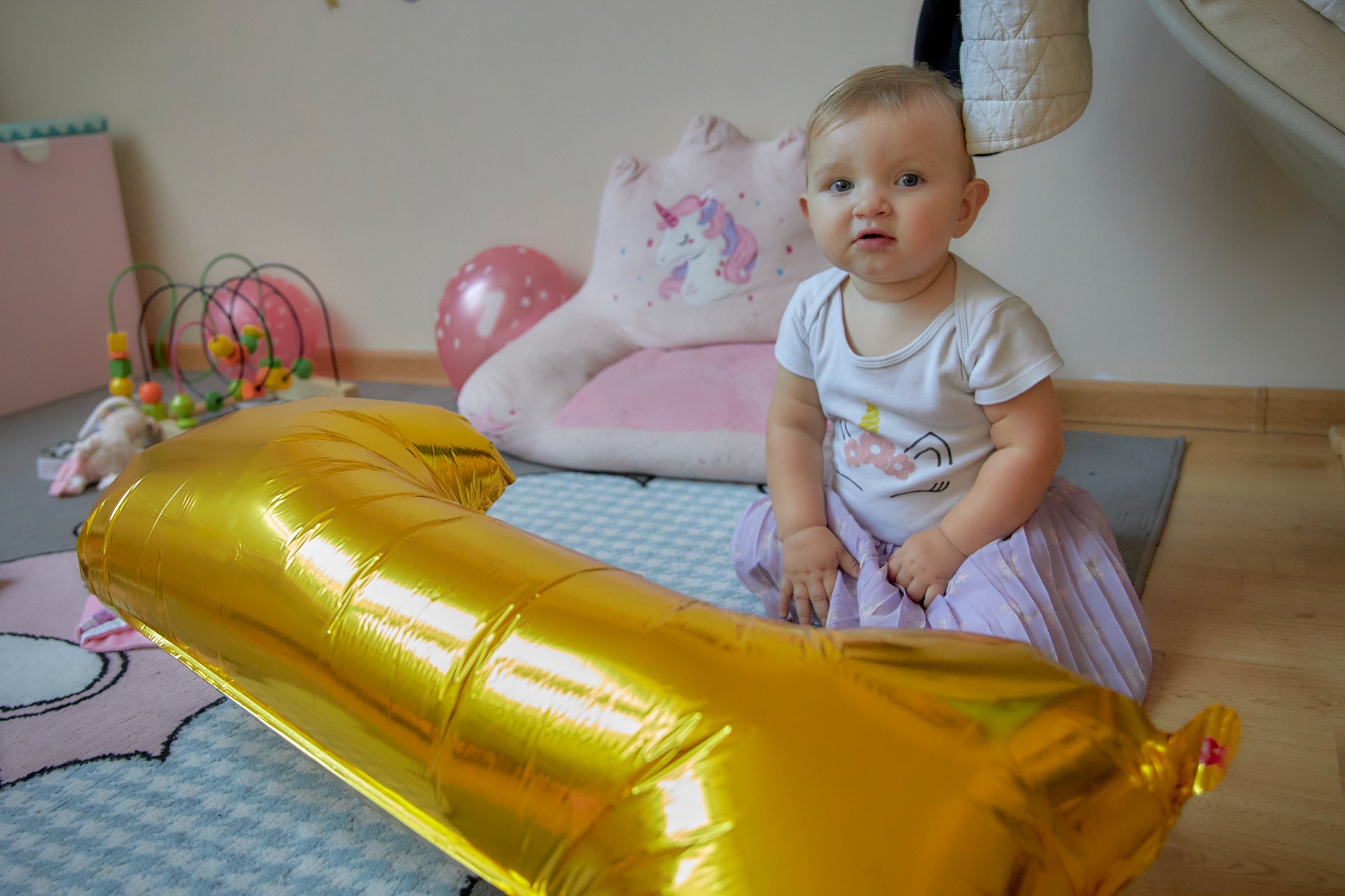 Roczek dziewczynki: roczne dziecko siedzi na dywanie obok dużego złotego balona w kształcie cyfry 1, w tle poduszka w kształcie jednorożca i inne zabawki.