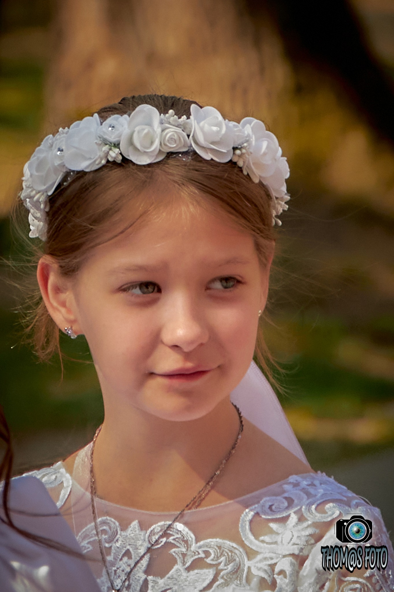 Portret dziewczynki w białej sukience i wianku z białych róż, z delikatnym uśmiechem i kolczykami w uszach, na rozmytym, naturalnym tle.