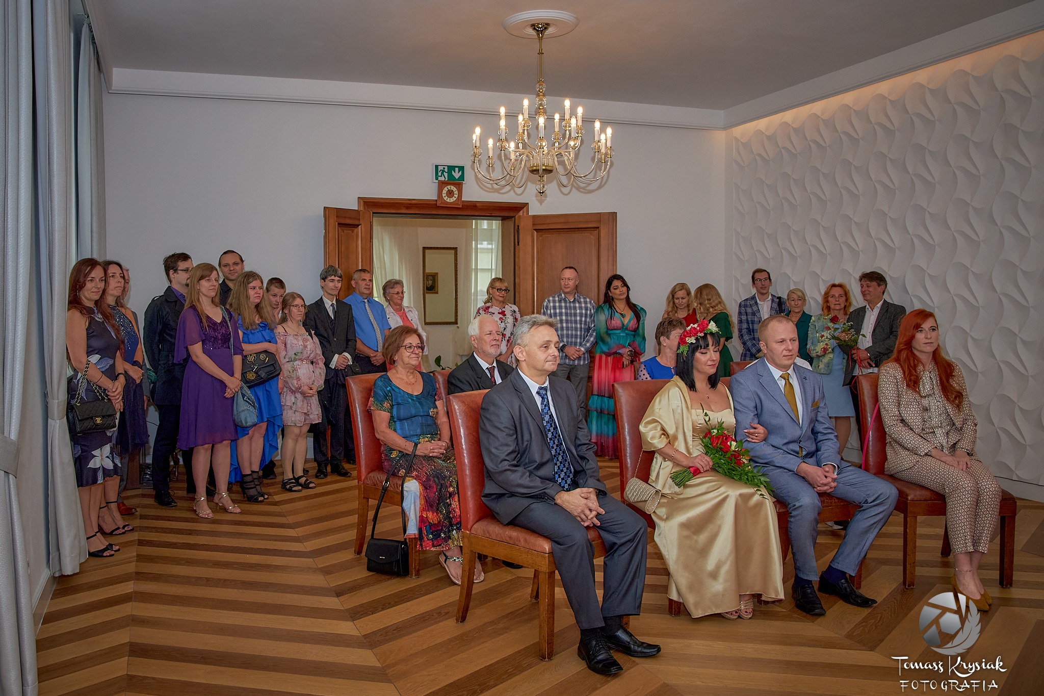 Uroczystość ślubna w urzędzie: Para młoda siedzi na krzesłach, trzymając bukiet czerwonych róż, otoczona gośćmi w eleganckich strojach, na tle białych ścian z teksturą i parkietu w jodełkę.