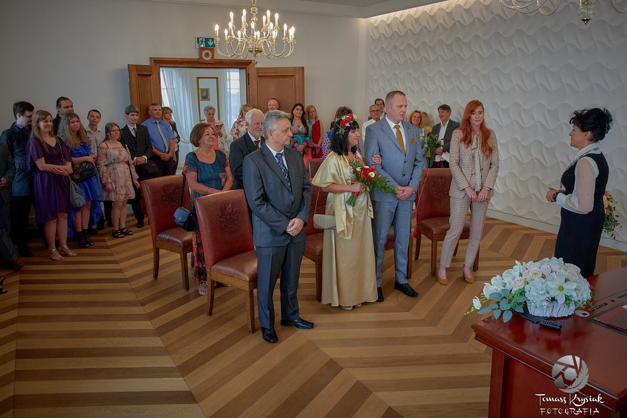 Para młoda podczas ceremonii ślubnej w urzędzie, z gośćmi w tle, na parkiecie w jodełkę, oświetlona żyrandolem.