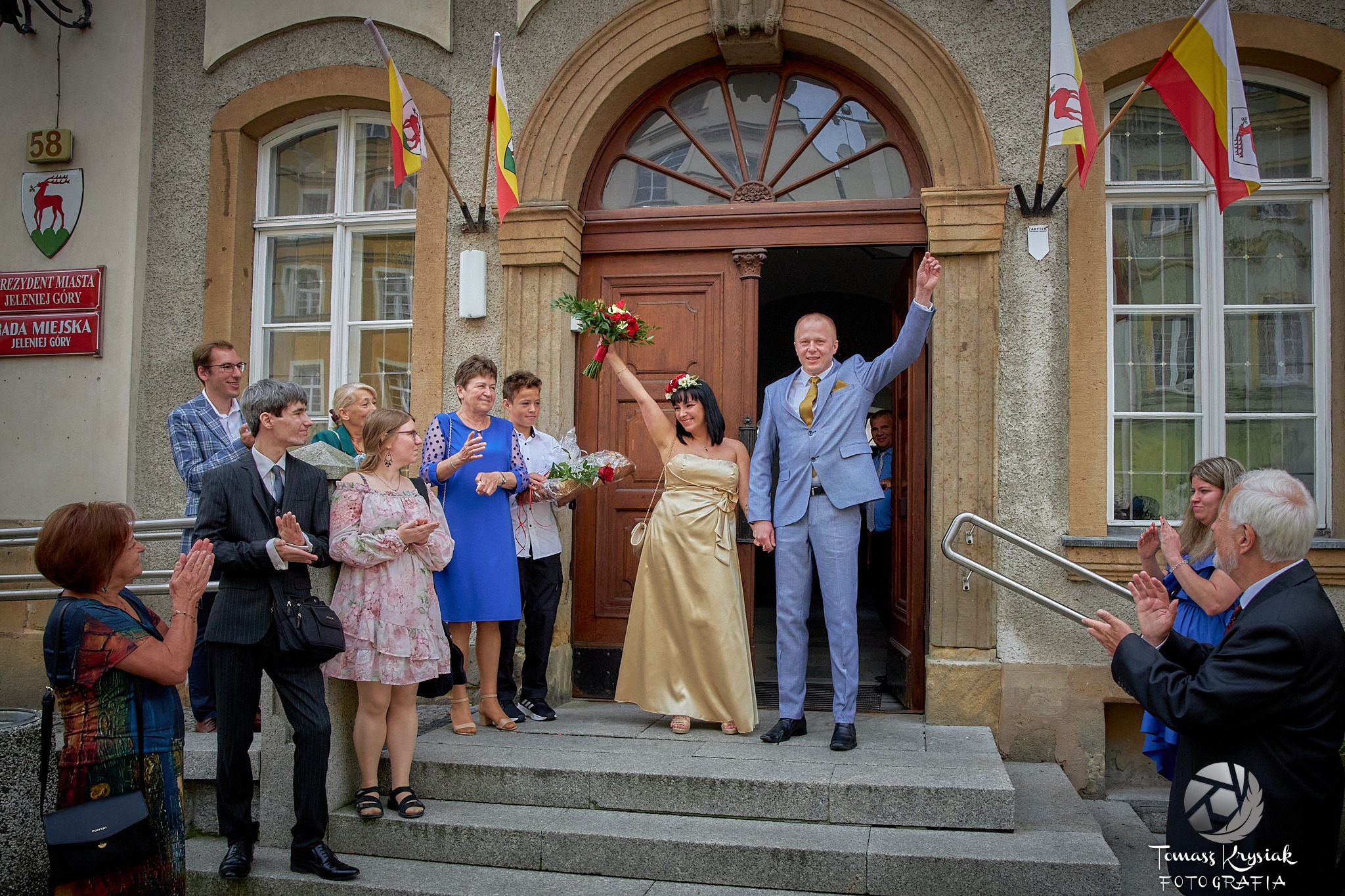 Para młoda w jasnych strojach, wychodząca z Urzędu Miasta Jeleniej Góry, witana oklaskami gości.