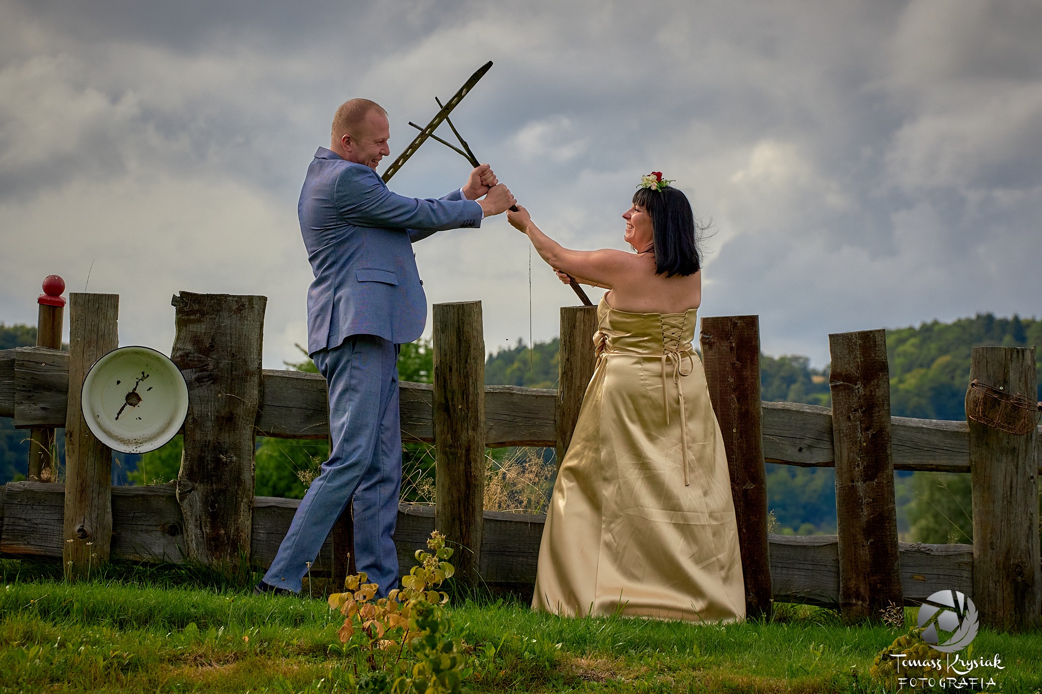 Para młoda w plenerze, przecinająca grabiami starą, drewnianą zagrodę; panna młoda w złotej sukni i z kwiatami we włosach, pan młody w niebieskim garniturze, oboje uśmiechnięci.