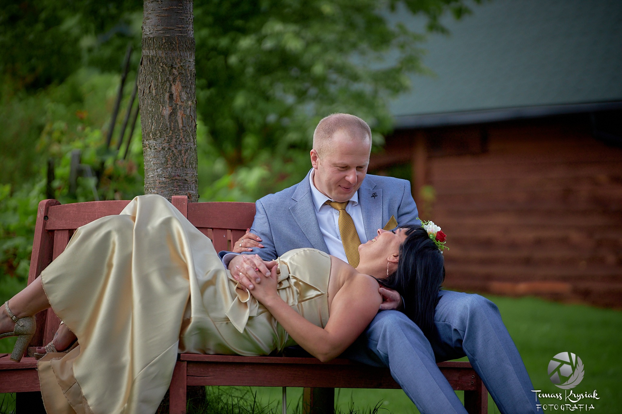 Para młoda siedząca na drewnianej ławce w parku, panna młoda w złotej sukni leży na kolanach pana młodego, oboje patrzą na siebie z uśmiechem.