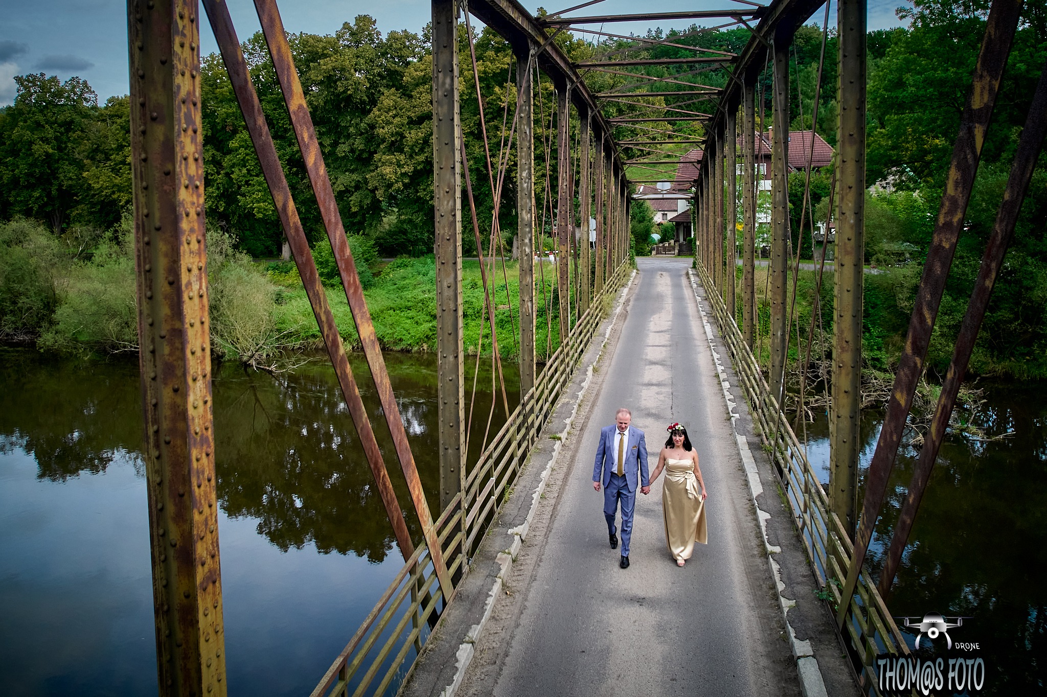 Para młoda, on w niebieskim garniturze, ona w złotej sukni, idąca po starym, metalowym moście z widokiem na rzekę i zieleń, ujęcie z drona.