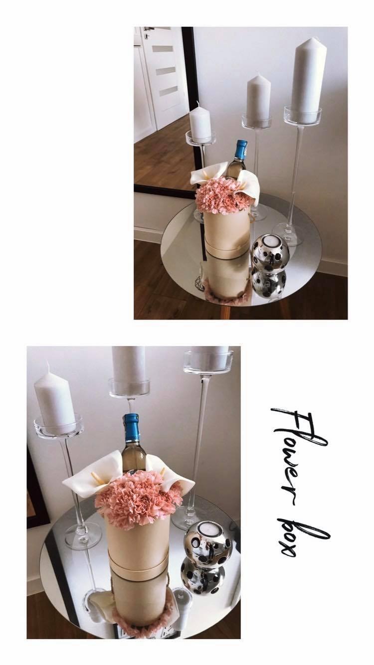 Dekoracja stołu: okrągły, lustrzany stolik z kompozycją kwiatową w beżowym pudełku, butelką z niebieską etykietą, białymi kaliami, dwoma srebrnymi świecznikami kulistymi i trzema wysokimi...