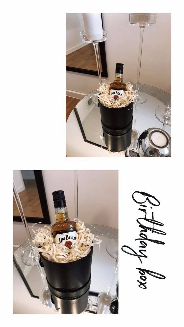 Prezent urodzinowy: butelka Jim Beam w czarnym pudełku wypełnionym jasnym siankiem na lustrzanej tacy, obok świecznika i kieliszków do wina.