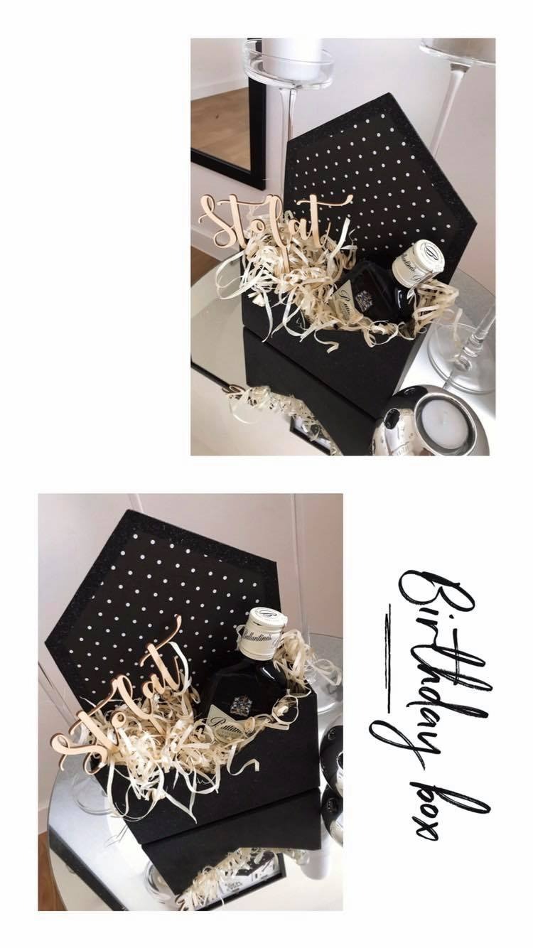 Zestaw prezentowy w czarnym pudełku z białymi kropkami, wypełniony ozdobnym siankiem, butelką alkoholu i drewnianym napisem 'Stolat', na szklanym stoliku z dekoracjami.