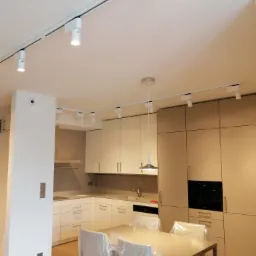 Nowoczesna kuchnia z beżowymi szafkami i białym systemem szynowym oświetlenia punktowego na suficie, z lampami skierowanymi w dół, dodatkowo lampa wisząca nad stołem. Krzesła zabezpieczone folią.