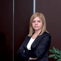 Portret kobiety w czarnej marynarce z założonymi rękami na tle ciemnej ściany i rośliny doniczkowej.