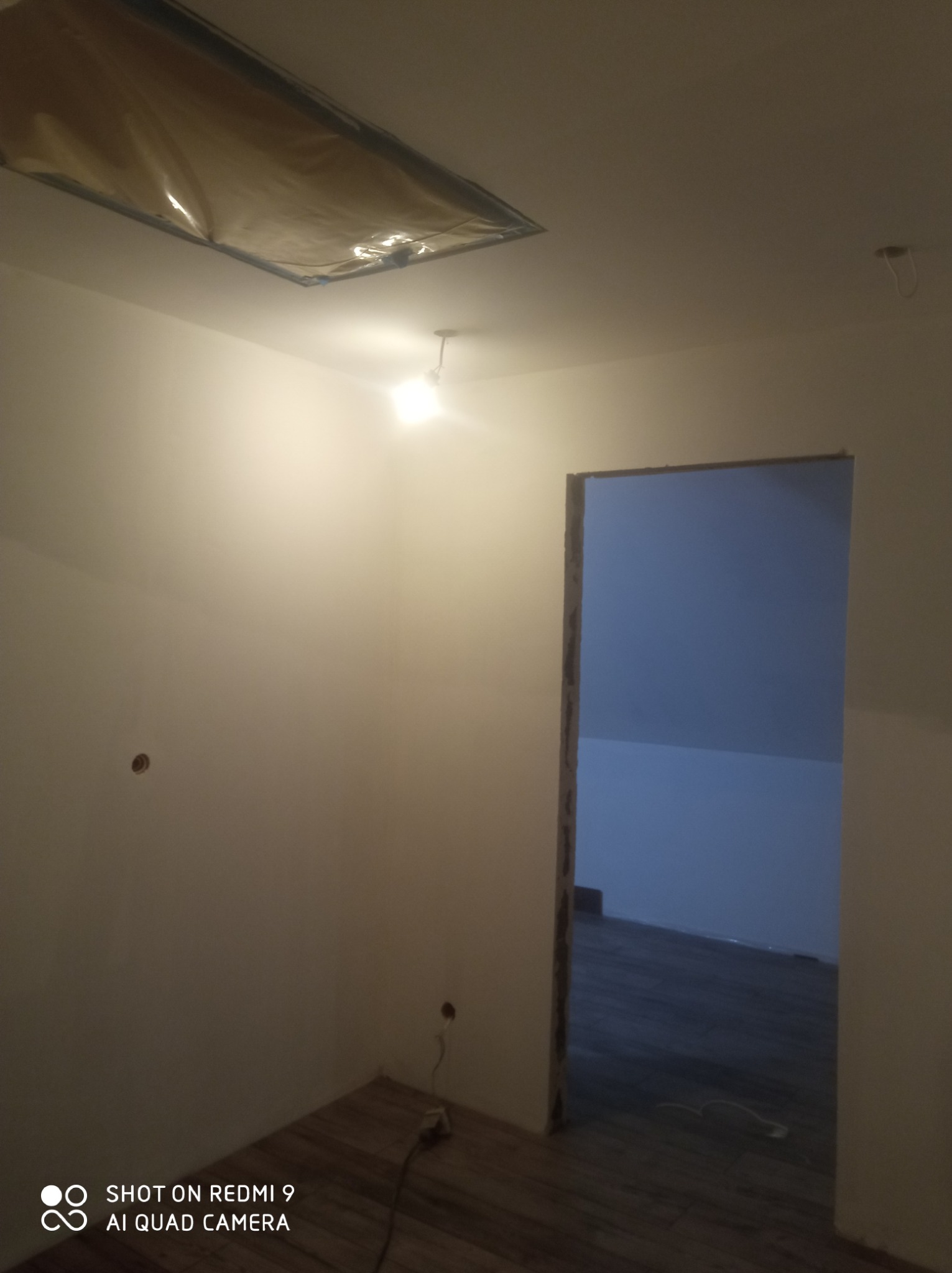 Pokój po szpachlowaniu ścian i sufitu, widoczne przygotowane otwory na gniazdka elektryczne, zabezpieczona folią część sufitu, oraz widok na poddasze z drewnianą podłogą przez otwór w ścianie.