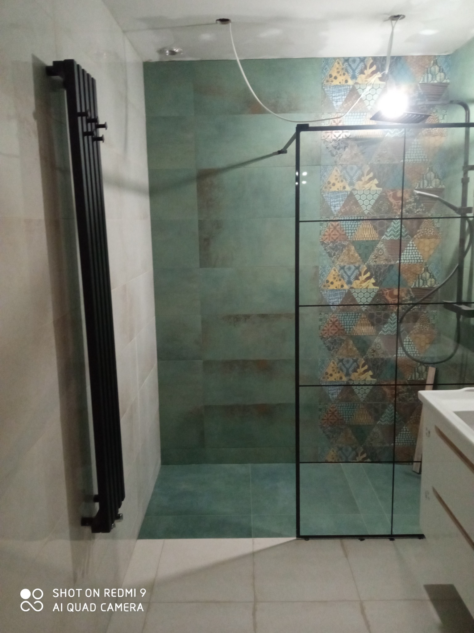 Wykończona łazienka z zielonymi płytkami i czarnym grzejnikiem drabinkowym, kabina prysznicowa z czarnymi profilami i wzorzystą ścianą w tle.