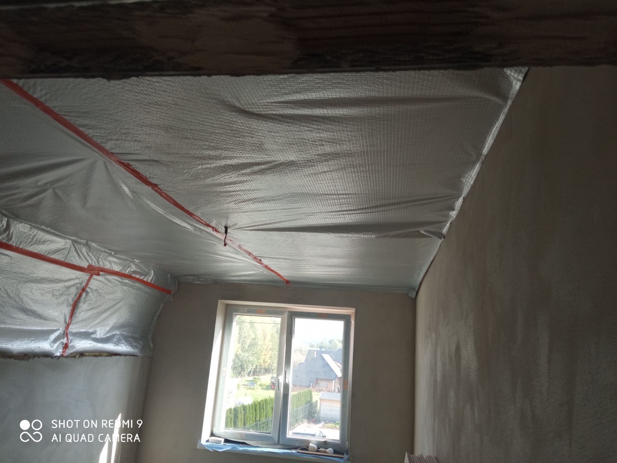 Montaż srebrnej folii termoizolacyjnej na poddaszu, zabezpieczonej czerwoną taśmą, widok na okno z widokiem na domy i zieleń.