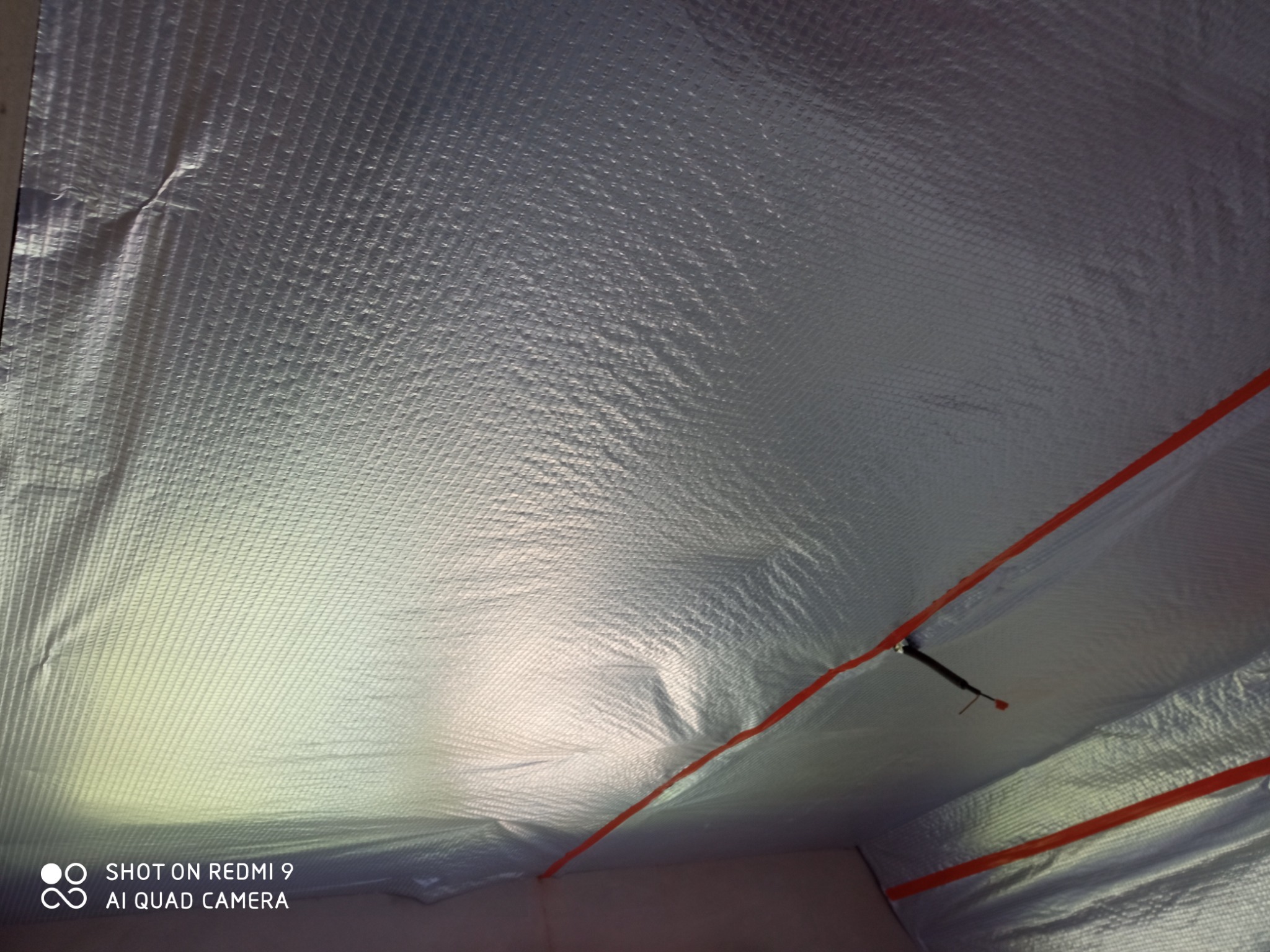 Izolacja poddasza folią aluminiową z widocznymi czerwonymi taśmami mocującymi i elementami konstrukcyjnymi, ujęcie z dołu.