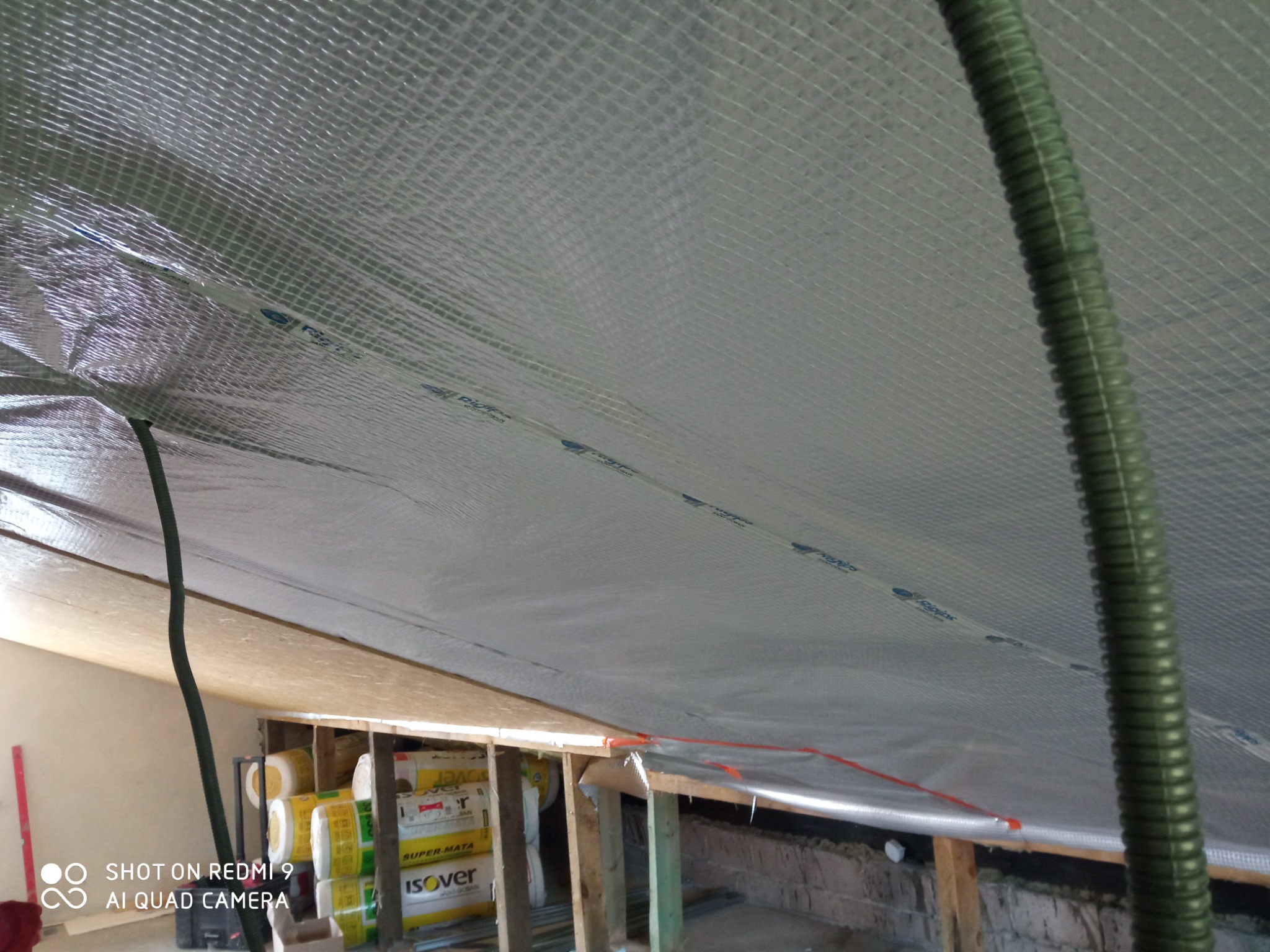 Izolacja poddasza z widoczną folią termoizolacyjną z nadrukiem, zielony wąż wentylacyjny i paczki wełny mineralnej Isover u dołu.