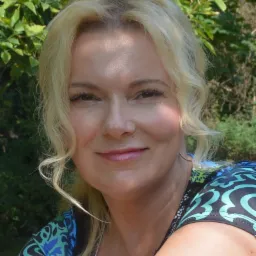 Portret uśmiechniętej kobiety z blond włosami na tle zielonej roślinności, ubrana w bluzkę z niebiesko-zielonym wzorem.