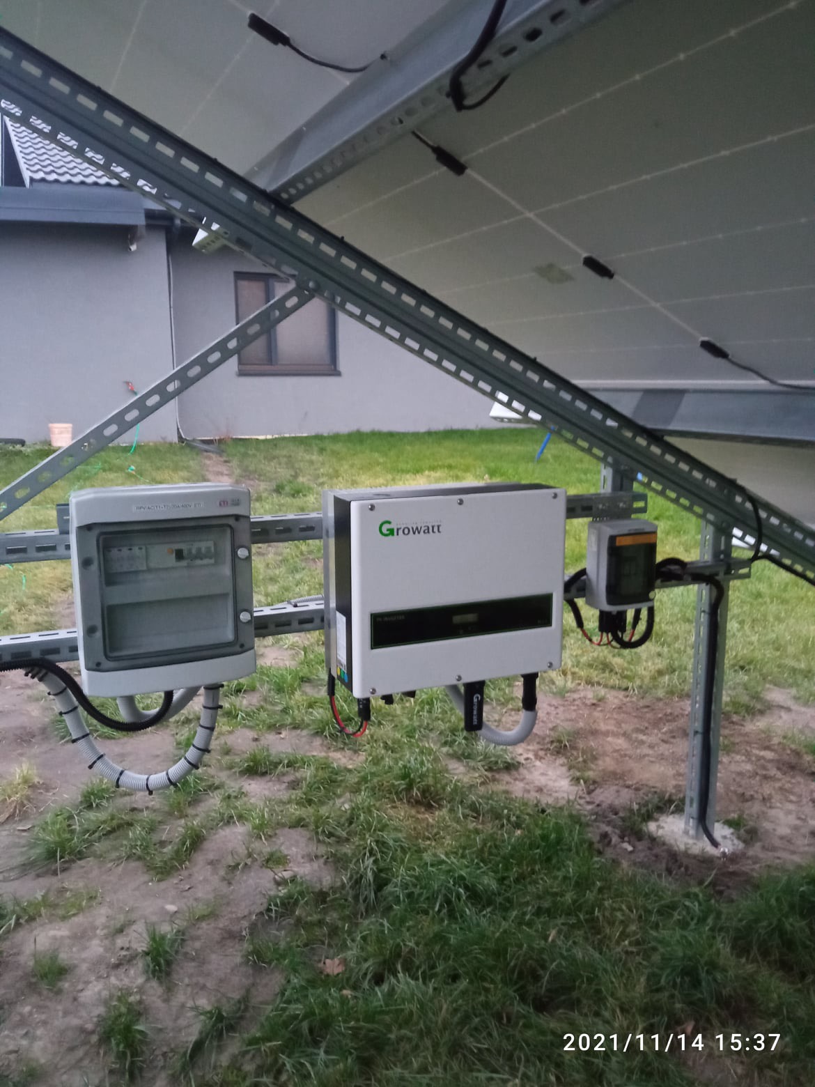 Instalacja z inwerterem Growatt i skrzynką zabezpieczeń zamontowana pod panelami słonecznymi na metalowej konstrukcji, widoczny fragment budynku mieszkalnego w tle.