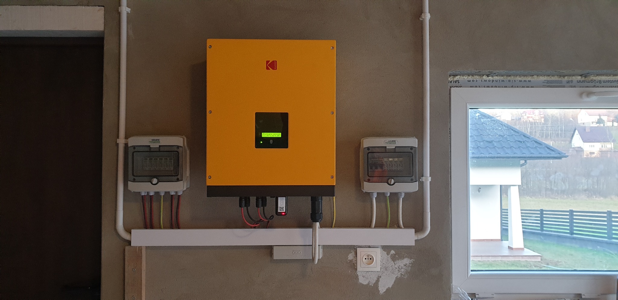 Żółty inwerter z logo na szarej ścianie wewnątrz budynku, po bokach dwie szare skrzynki elektryczne, widoczne kable i przewody, w tle okno z widokiem na domy i zieleń.