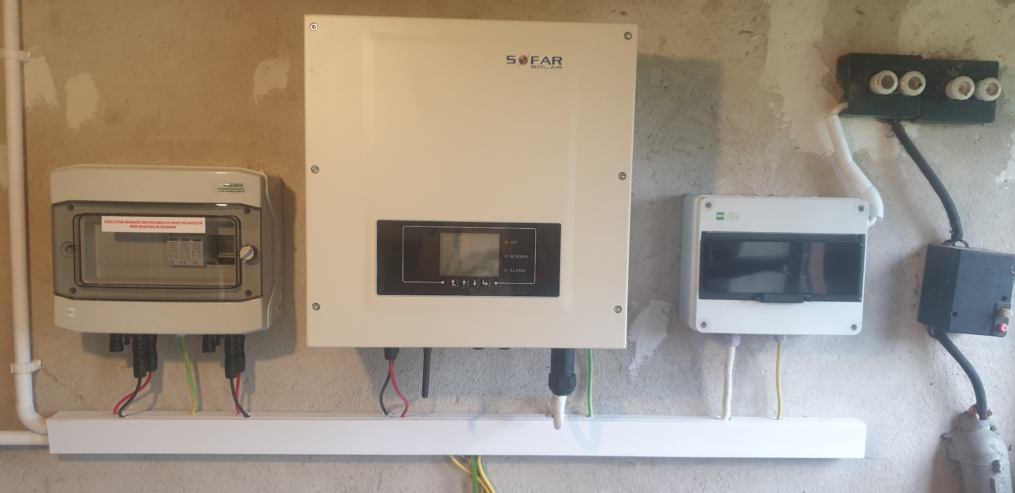 Biały inwerter SOFAR SOLAR zamontowany na szarej ścianie obok skrzynki z bezpiecznikami i rozdzielnicy elektrycznej, widoczne przewody i rurki instalacyjne.