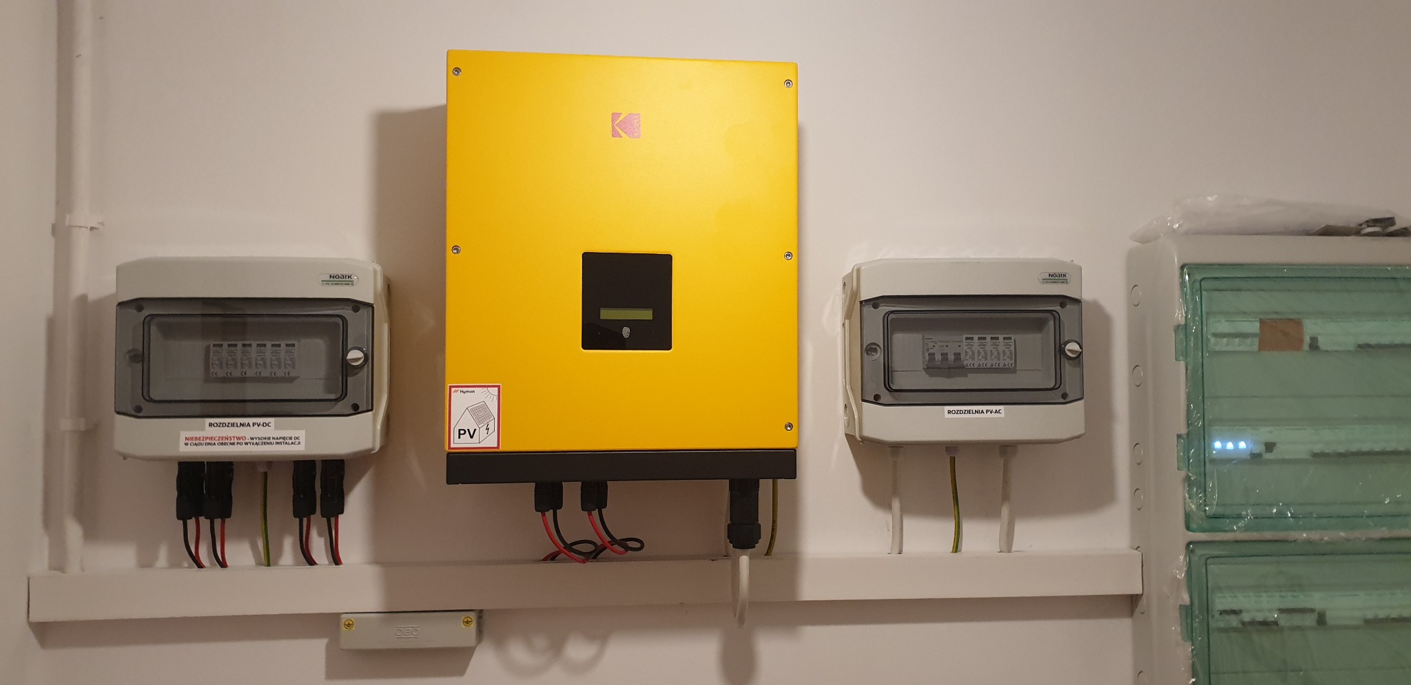 Żółty falownik z logiem K oraz rozdzielnice PV-DC i PV-AC zamontowane na białej ścianie, widoczne przewody i naklejka informacyjna.