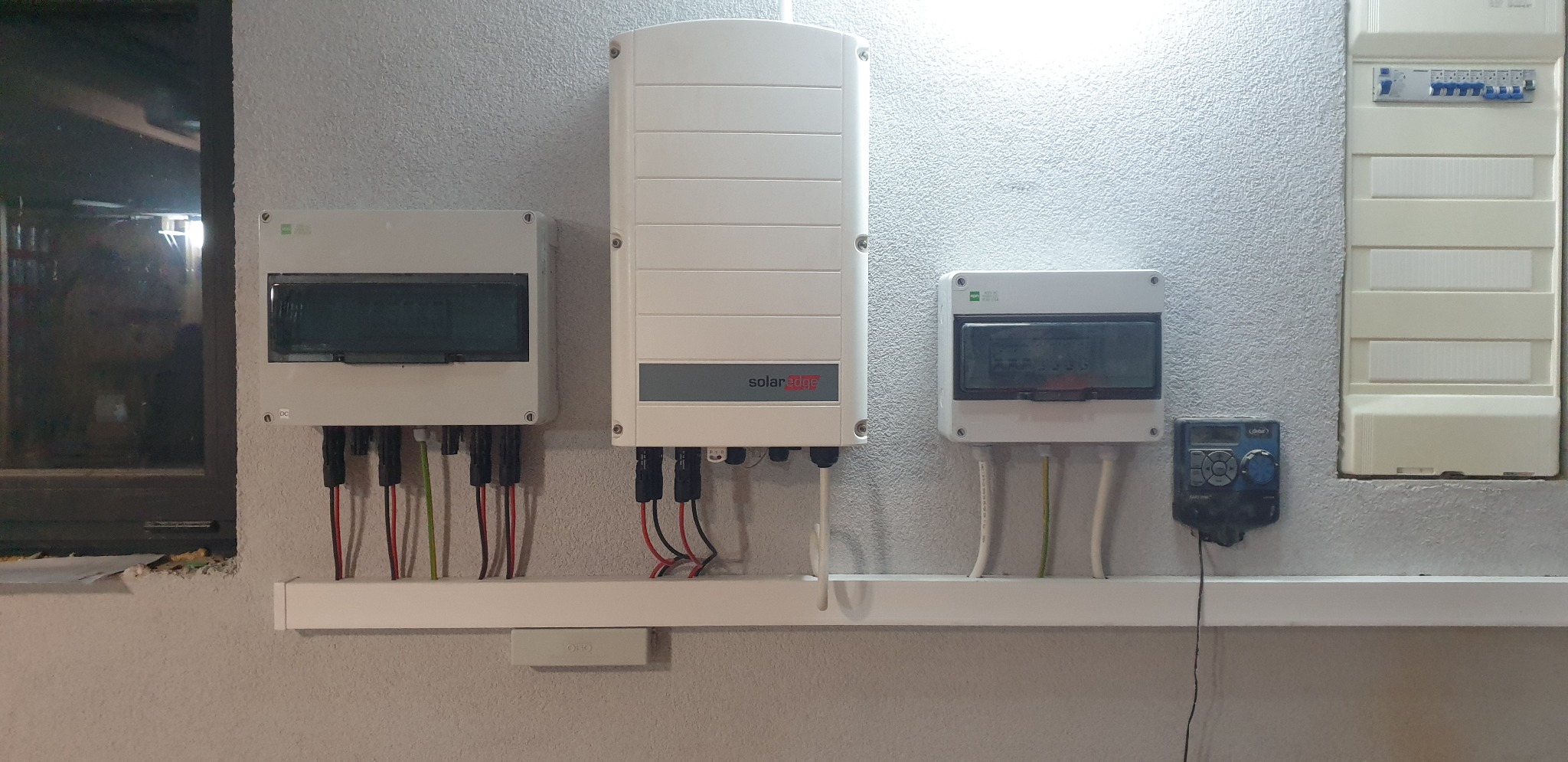 Instalacja elektryczna z falownikiem SolarEdge i skrzynkami rozdzielczymi na szarej, teksturowanej ścianie wewnątrz budynku.
