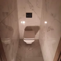 Wąska toaleta w wykończeniu z białych płytek imitujących marmur, z zamontowaną podwieszaną miską WC z szarą deską i czarnym przyciskiem spłukującym.