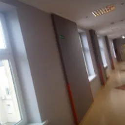 Korytarz biurowy z pomalowanymi ścianami na szaro, widoczne okna z białymi ramami, oświetlenie sufitowe i jasna podłoga.