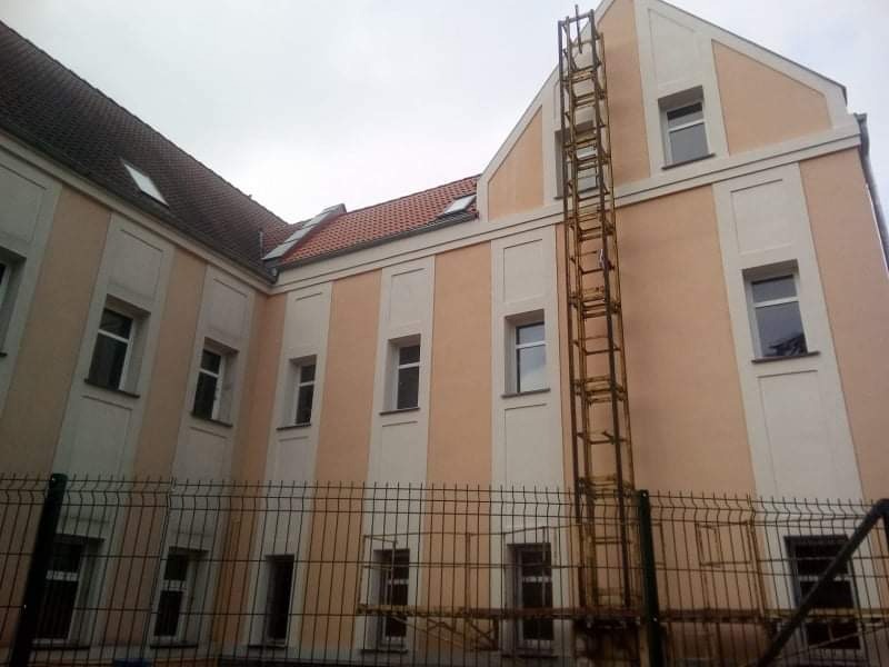 Odnowiona fasada budynku z elementami dekoracyjnymi, widoczna metalowa konstrukcja rusztowania przy ścianie, ogrodzenie z siatki na pierwszym planie.