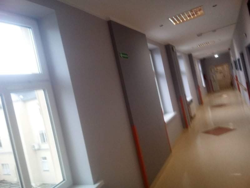 Korytarz biurowy z pomalowanymi ścianami na szaro, widoczne okna z białymi ramami, oświetlenie sufitowe i jasna podłoga.