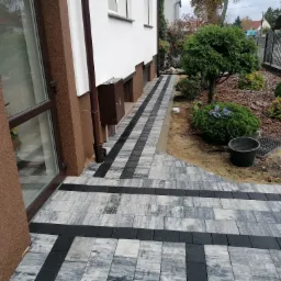 Ścieżka z kostki brukowej w odcieniach szarości i czerni prowadząca wzdłuż brązowej ściany budynku i zielonego ogrodu z drzewami i krzewami, widoczny czarny kubeł i metalowa skrzynka na listy.