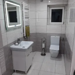 Wykończona łazienka w szaro-białej kolorystyce z podświetlanym lustrem, grzejnikiem drabinkowym z powieszonym ręcznikiem i toaletą.