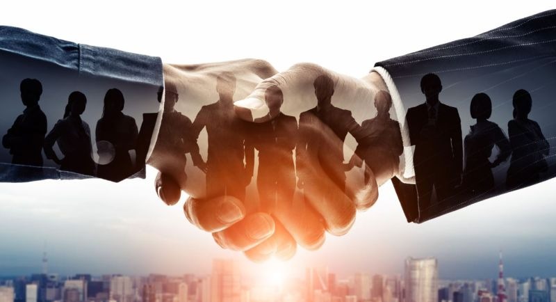Uścisk dłoni dwóch osób na tle panoramy miasta, z nałożonymi sylwetkami grupy ludzi, sugerujący współpracę i partnerstwo.