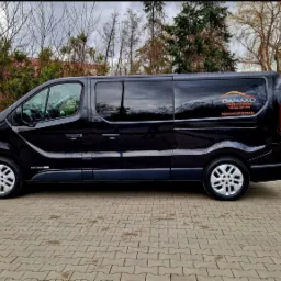 Czarny bus firmy przewozowej Damako z naklejonym logo i danymi kontaktowymi, zaparkowany na brukowanym placu, na tle zieleni.