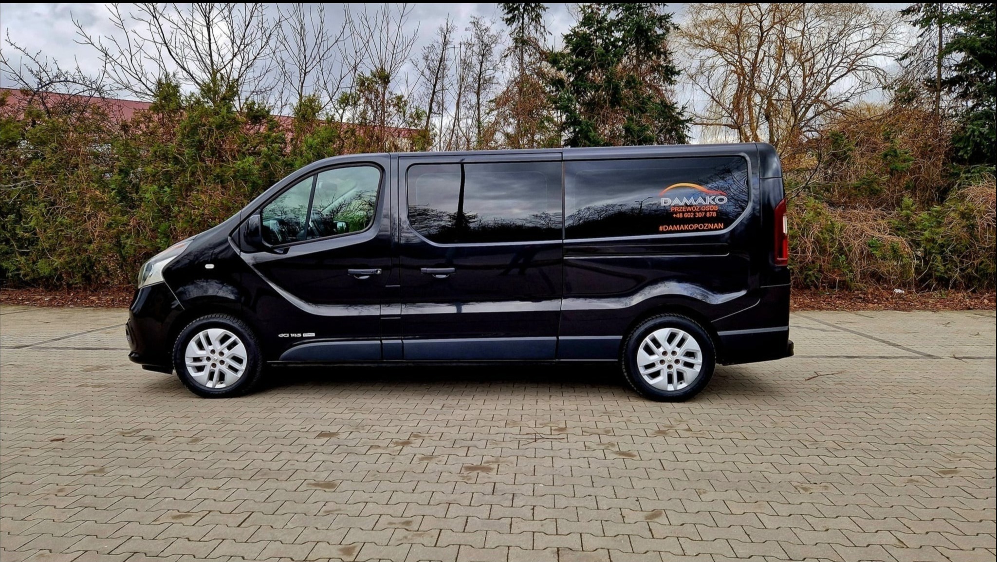 Czarny bus firmy przewozowej Damako z naklejonym logo i danymi kontaktowymi, zaparkowany na brukowanym placu, na tle zieleni.