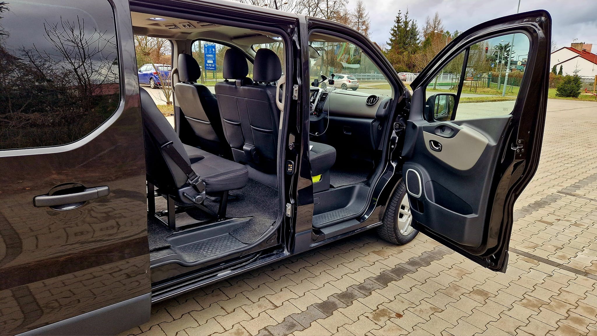 Czarny van z otwartymi drzwiami bocznymi i widocznym wnętrzem z czarnymi siedzeniami i pasami bezpieczeństwa, na zewnątrz kostka brukowa i drzewa.