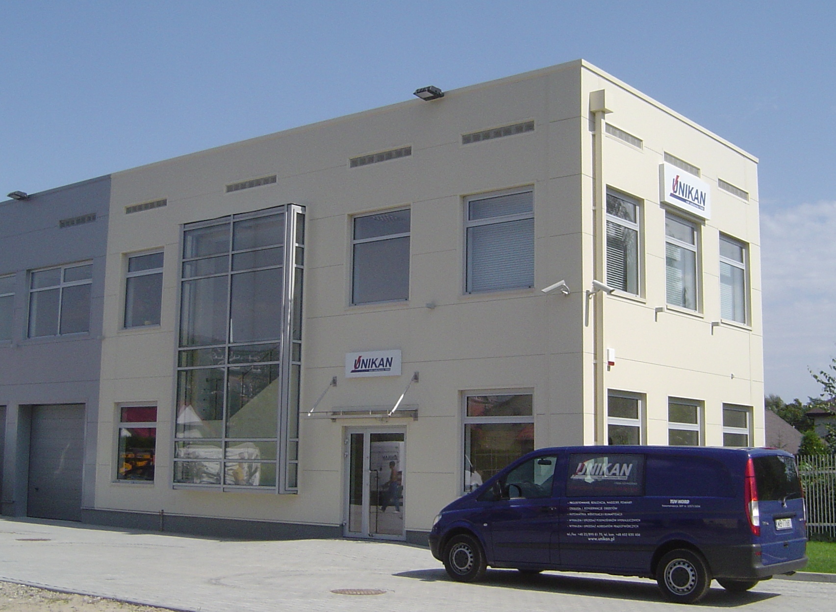 Dwupiętrowy budynek biurowy z dużymi oknami i ciemnoniebieskim samochodem dostawczym z logo firmy, widoczne kamery monitoringu i oświetlenie zewnętrzne.