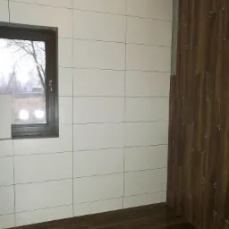 Wnętrze łazienki w trakcie układania płytek: białe płytki na ścianie z oknem i brązowe płytki imitujące drewno na sąsiedniej ścianie, widoczne krzyżyki dystansowe.