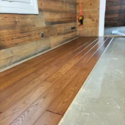 Układanie drewnianej podłogi z widocznym laserem poziomującym na ścianie z boazerii, część podłogi jeszcze nieukończona.