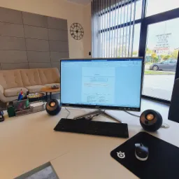 Biuro z biurkiem, komputerem z otwartym dokumentem tekstowym zawierającym wizualizację domu, głośnikami, myszką na podkładce, oraz sofą w tle. Na ścianie widoczny zegar.