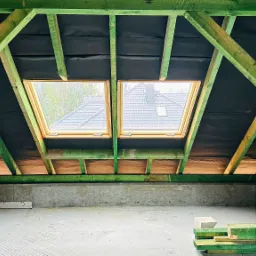 Konstrukcja dachu z zielonych belek drewnianych, widoczne dwa okna dachowe z widokiem na dom i drzewa, czarna membrana dachowa, surowy betonowy strop.