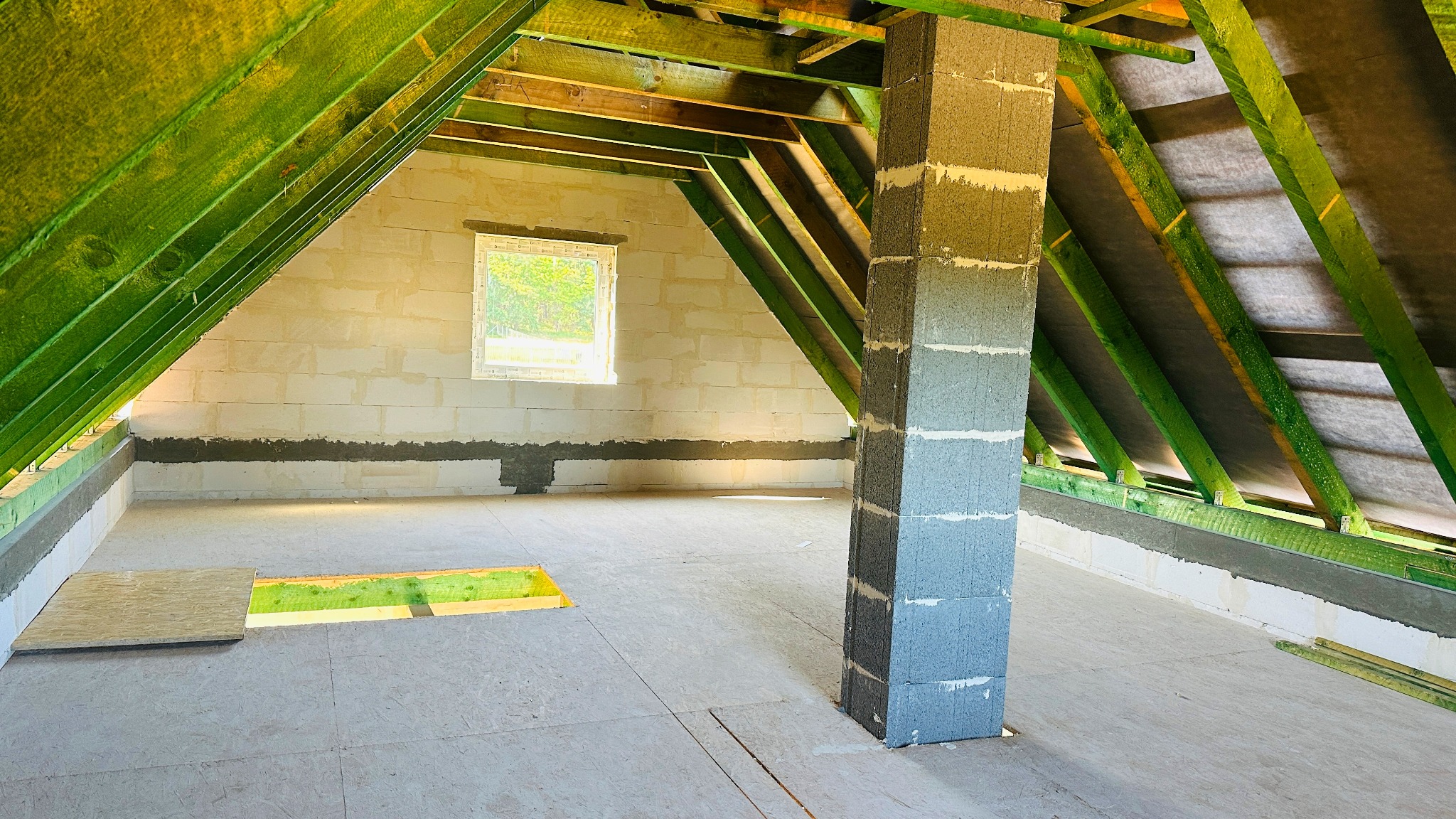 Wnętrze poddasza w trakcie budowy, widoczne ściany z betonu komórkowego, drewniana konstrukcja dachu z zieloną impregnacją oraz częściowo ułożona podłoga z płyt OSB, z otworem na schody.