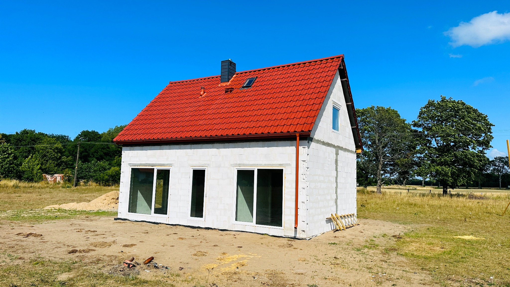 Niedokończony, parterowy budynek z białych bloczków z czerwoną dachówką na tle błękitnego nieba i zieleni. Widoczne okna bez szyb, drabina oparta o ścianę i stos piasku w tle.