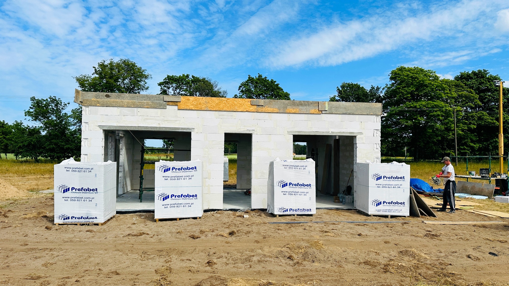 Wznoszenie surowego stanu budynku z białych bloczków, widoczne palety z materiałem budowlanym firmy Prefabet oraz pracownik z piłą tarczową w tle.
