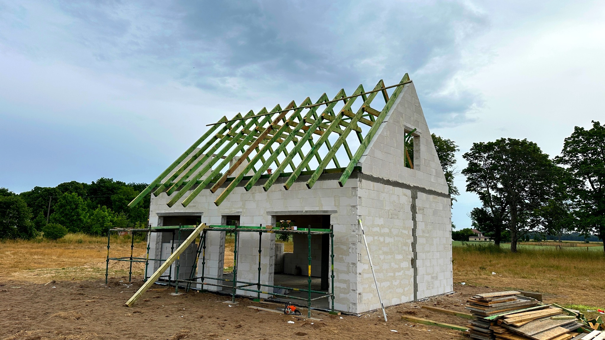 Surowy stan domu z białych bloczków, widoczna konstrukcja dachu z zielonych belek, rusztowania i deski leżące na ziemi, w tle łąka i drzewa.