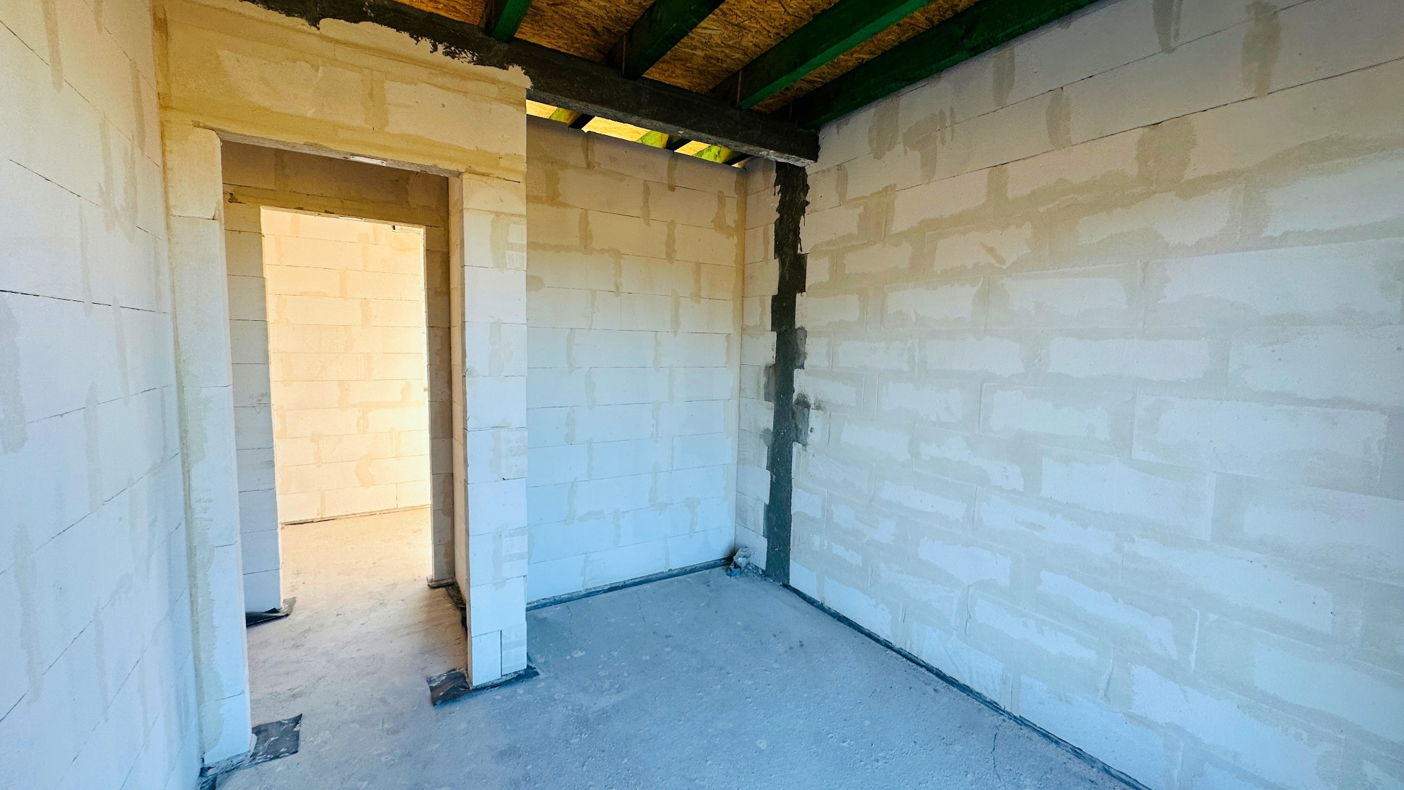 Wnętrze w stanie surowym z białych bloczków, widoczne ślady zaprawy, sufit z drewnianych belek pomalowanych na zielono, widok na kolejne pomieszczenie przez otwór drzwiowy.