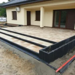 Wykończony taras z kostki brukowej, prowadzący do domu, z trzema stopniami i obrzeżami z ciemnych bloczk&oacute;w. W tle pracownik z taczką i palety z materiałem.