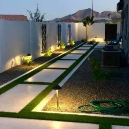 Oświetlona ścieżka z płyt betonowych rozdzielonych sztuczną trawą, prowadząca przez żwirowe podwórko, z dekoracyjnymi elementami na białym murze i lampkami w tle.