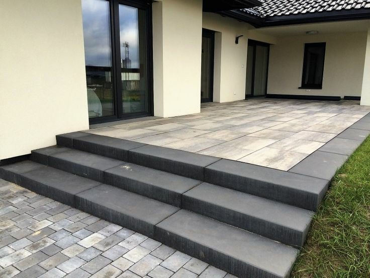 Nowoczesny taras z szarych płyt betonowych, schody z ciemnoszarej kostki, widok na elewację domu z dużymi oknami.